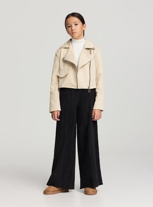 Giacca corta beige con chiusura a zip e tasca laterale, abbinata a pantaloni neri a gamba larga e calzature color cuoio. Tessuto dalla texture liscia.