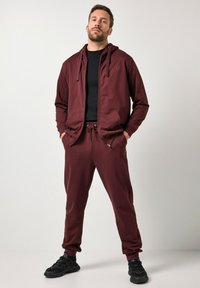 Sudadera con cremallera en color marrón granate, con cordones y bolsillos frontales, combinada con joggers ajustados a juego. Camiseta negra debajo, acompañada de zapatillas negras.