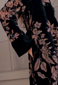 Juodo aksomo rūbas, puoštas išsamiais smėlio spalvos gėlių siuvinėjimais ant rankovių ir kūno. Dizainą sudaro lapai ir žiedai.