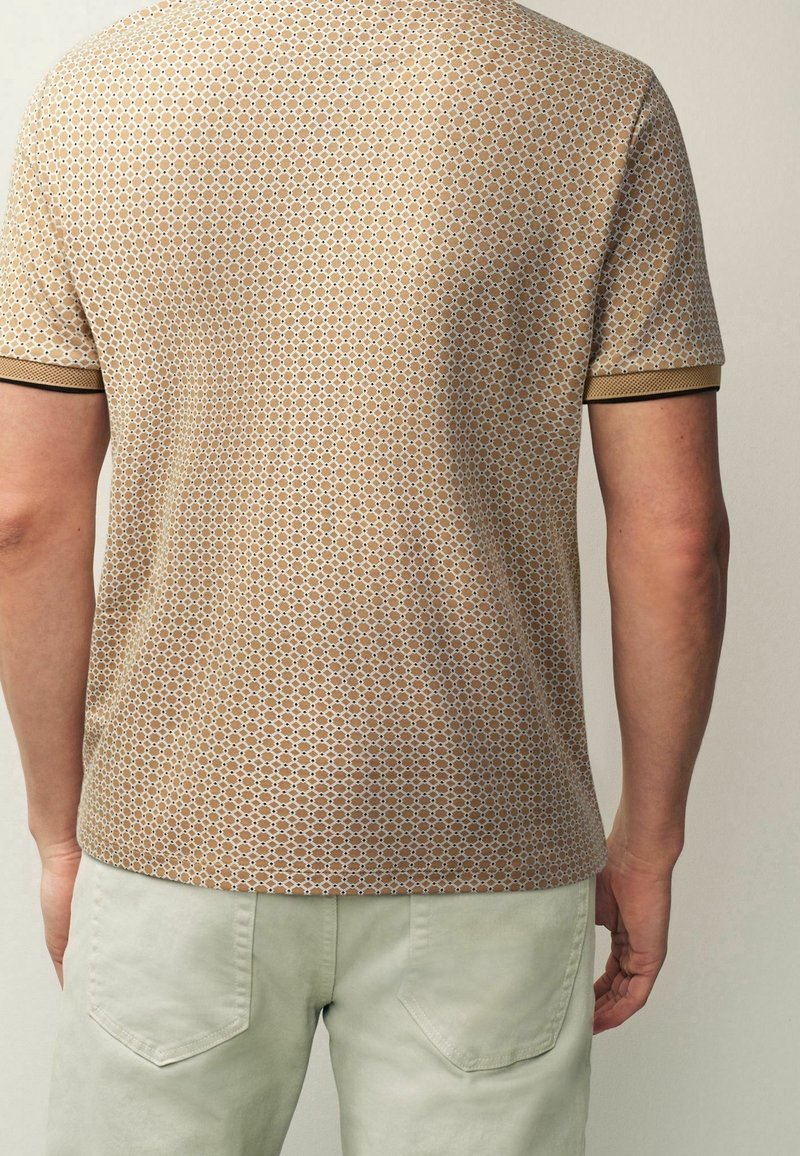 Homme portant une chemise beige à manches courtes à motifs et un pantalon de couleur claire, vu de dos sur un fond uni.