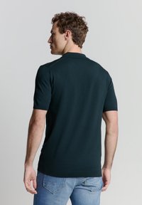 Donkergroene polo met korte mouwen, geribde kraag en textuurstof. Van achterkant bekeken, gecombineerd met lichtblauwe jeans.