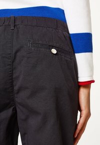 Pantalon noir avec une poche latérale et une fermeture à bouton. Tissu doux et texturé avec une coupe décontractée. Pull rayé bleu et blanc visible.