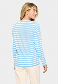 Vrouw die glimlacht en over haar schouder kijkt, gekleed in een lichtblauwe en witte gestreepte lange mouwen shirt met crèmekleurige broek op een witte achtergrond.