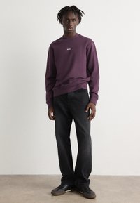 Sudadera morada con el logo blanco "BOSS", cuello redondo, puños de canalé, combinada con jeans negros y zapatillas oscuras sobre un fondo neutro.