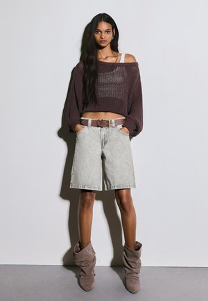 Žena s dlhými tmavými vlasmi má na sebe voľný hnedý pletený sveter, sivé šortky po kolená s opaskom a voľné taupe čižmy, stojaca pri jednoduchej stene.