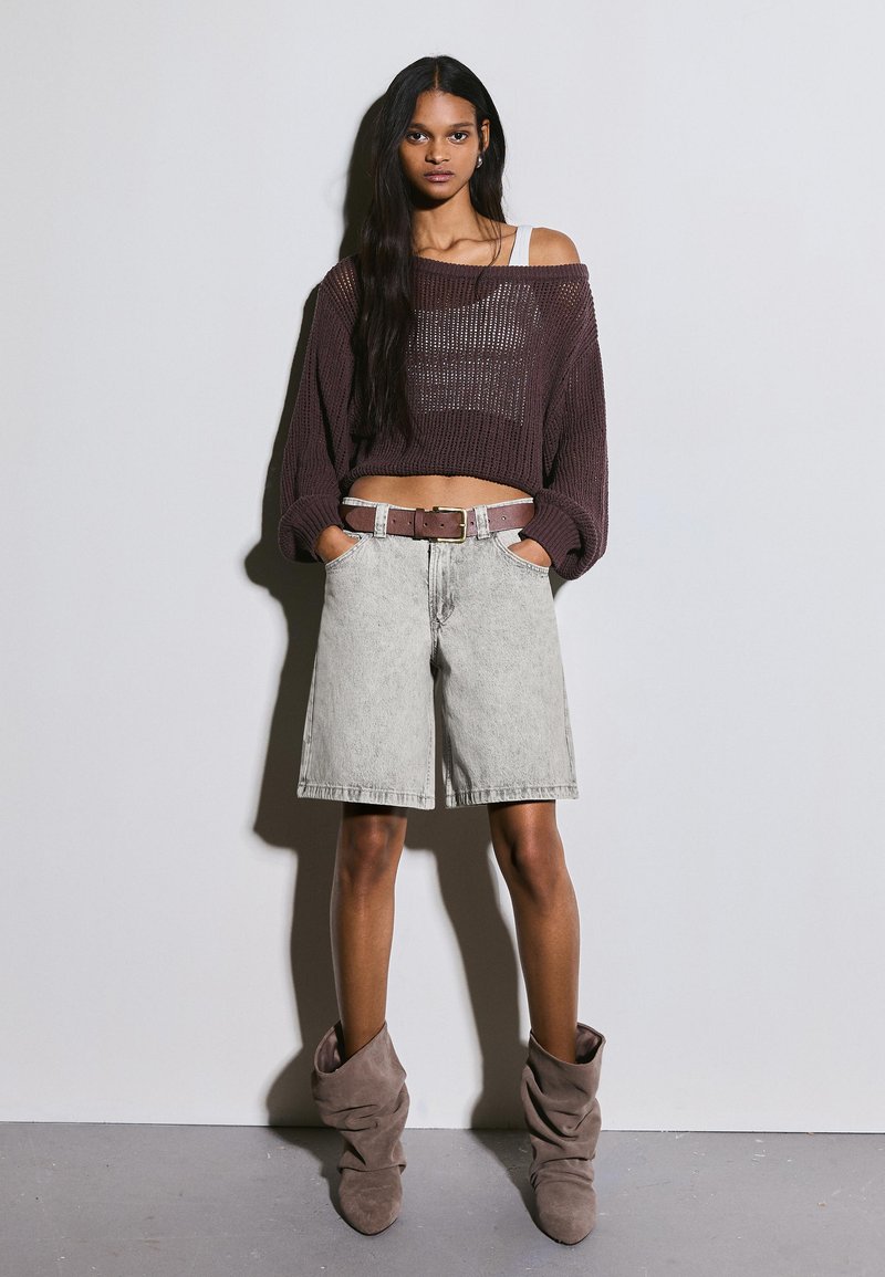 Femeie cu păr lung și închis la culoare poartă un pulover maro lejer tricotat, pantaloni scurți gri cu centură până la genunchi și cizme largi de culoare taupe, stând în picioare în fața unui perete simplu.