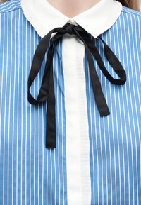 Μπλούζα με κάθετες ρίγες σε μπλε και άσπρο, με λευκή γιακά με βολάν και μαύρη υφασμάτινη φιόγκο δεμένη στο λαιμό.