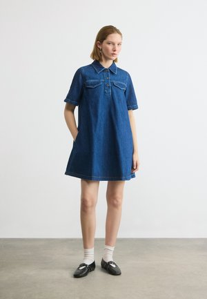 Femeie în picioare, purtând o rochie scurtă din denim albastru cu buzunare în față, șosete albe și balerini negri, pe un podea gri.