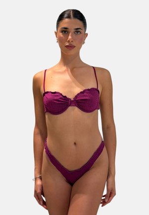 Giovane donna con capelli scuri raccolti indossa un bikini viola a due pezzi con bordi smerlati, in piedi davanti a uno sfondo chiaro e uniforme.