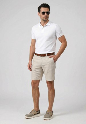 Mann trägt weißes Polohemd, beige Shorts mit braunem Gürtel, graue Sneaker und Sonnenbrille, steht mit einer Hand in der Tasche.