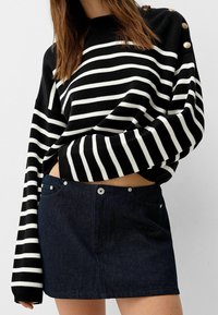 Pull rayé noir et blanc avec des manches surdimensionnées et des accents de boutons dorés, associé à une mini-jupe en denim foncé ornée de clous argentés.