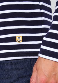 Stribet langærmet skjorte i navy og hvid lavet af bomuld, med et lille gult ankerlogo-patch. Teksturen er glat og tætsiddende.