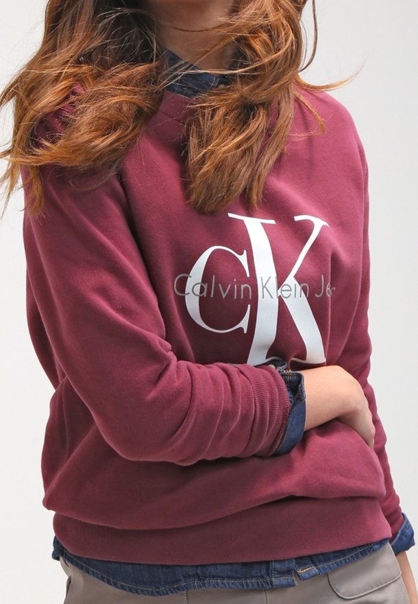 Sudadera burdeos con logo "CK" en blanco. Presenta un corte relajado con puños de canalé. Usada sobre una camisa de mezclilla. Tejido suave y texturizado.