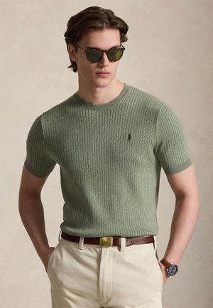 Mand iført grøn ribbet kortærmet sweater, beige bukser, brun bælte, håndledsur og mørke solbriller, stående med hænderne i lommerne.