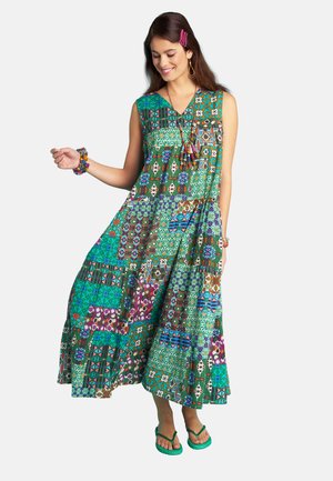 Young woman wearing a colorful sleeveless patchwork dress and green flip-flops, smiling and holding the dress out with one hand.