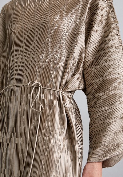 Close-up of taupe pleated dress with textured vertical lines and a thin waist tie knot on a person's arm and torso.