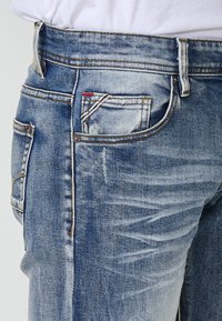 Jeans de denim azul con un acabado desgastado, cinco bolsillos, costuras en contraste y una pequeña etiqueta roja y blanca en el bolsillo delantero.
