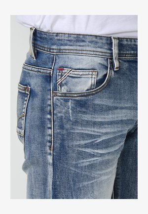 Jeans in denim blu con una finitura sbiadita, cinque tasche, cuciture a contrasto e una piccola etichetta rossa e bianca sulla tasca anteriore.