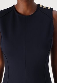 Granatowa, dopasowana sukienka bez rękawów z okrągłym dekoltem i trzema złotymi guzikami na ramionach. Gładki, elastyczny materiał z widocznymi szwami.