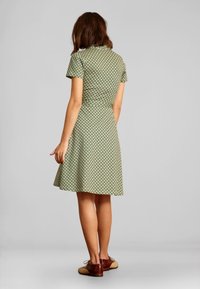 Zielona sukienka z krótkimi rękawami, dopasowanym gorsetem i rozkloszowaną spódnicą, zdobiona geometrycznym wzorem w jasnym niebieskim i brązowym kolorze. Brązowe buty z akcentami.