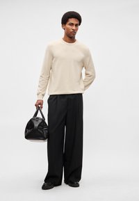 Jersey beige de punto, pantalones negros de pierna ancha, sosteniendo un bolso negro con textura. Hombre de pie con un fondo neutro.