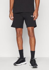 Férfi fekete sport rövidnadrágban, fekete sportzokniban és fekete sportcipőben áll világos padlón fehér fal előtt.