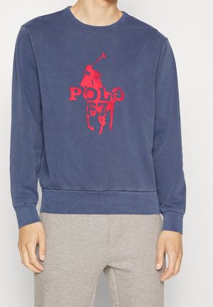 Sweat-shirt bleu marine avec graphisme de joueur de polo rouge et texte « POLO 67 », porté avec un pantalon gris clair.
