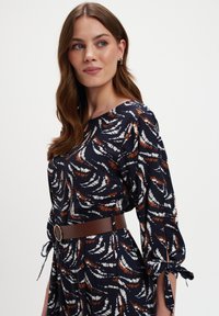 Kobieta z długimi brązowymi włosami, ubrana w sukienkę w granatowo-białe wzory z brązowymi akcentami, z rękawami o długości 3/4, związana u mankietów i szerokim brązowym paskiem.