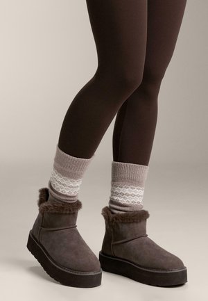 Barna szarvasbőr bokacsizma műszőrme szegéllyel, világosszürke mintás zoknival párosítva. A lábak barnás leggingsben vannak.