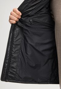 Una mano tiene aperto un giubbotto in pelle nera, mostrando una fodera interna nera liscia con una tasca con zip.