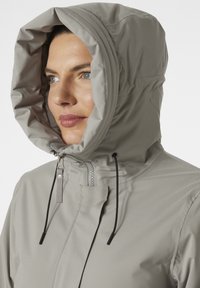 Svetle šedá voděodolná bunda s kapucí, vybavená předním zipem, stahovacími šňůrkami a hladkou texturou. Navržena pro funkčnost a pohodlí.
