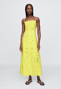 Gul maxikjole med tætsiddende overdel, tynde stropper og blomsterudskæringsmønstre. Bomuldsstof med glat tekstur, perfekt til sommer.