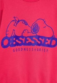 Dessin animé de Snoopy endormi en bleu sur tissu rouge au-dessus du mot « OBSESSED » et texte plus petit « GOODNESS & GRIEF. »