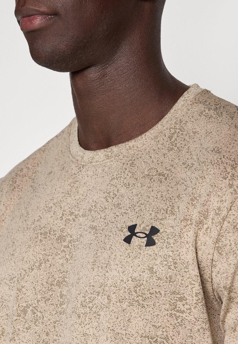 Primer plano de una persona que lleva una camiseta de Under Armour beige con motas y un logo negro en el pecho, sobre un fondo liso.