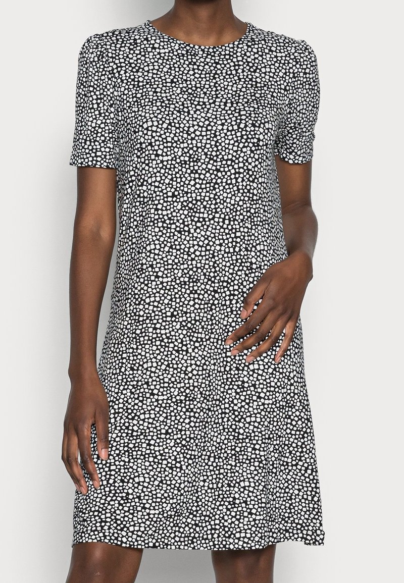 Femme portant une robe noire à manches courtes avec un motif de points blancs irréguliers, debout devant un fond clair uni.
