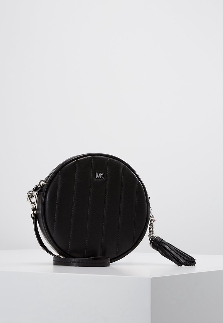 Polsino in pelle nera rotondo con cuciture verticali, logo MK argentato e tirazip a frange su un piedistallo bianco.