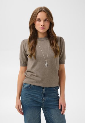 Mujer con cabello largo y castaño lleva un top de punto de manga corta en color topo, vaqueros azules y un collar largo de plata con colgante sobre un fondo liso.
