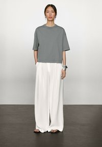 T-shirt gris court à manches courtes, associé à un pantalon blanc à jambes larges et des sandales plates noires. Accessoire : bracelet en argent.