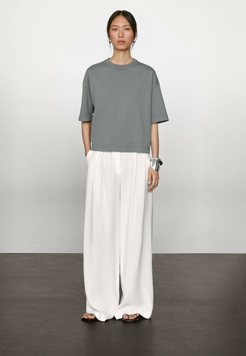T-shirt gris court à manches courtes, associé à un pantalon blanc à jambes larges et des sandales plates noires. Accessoire : bracelet en argent.