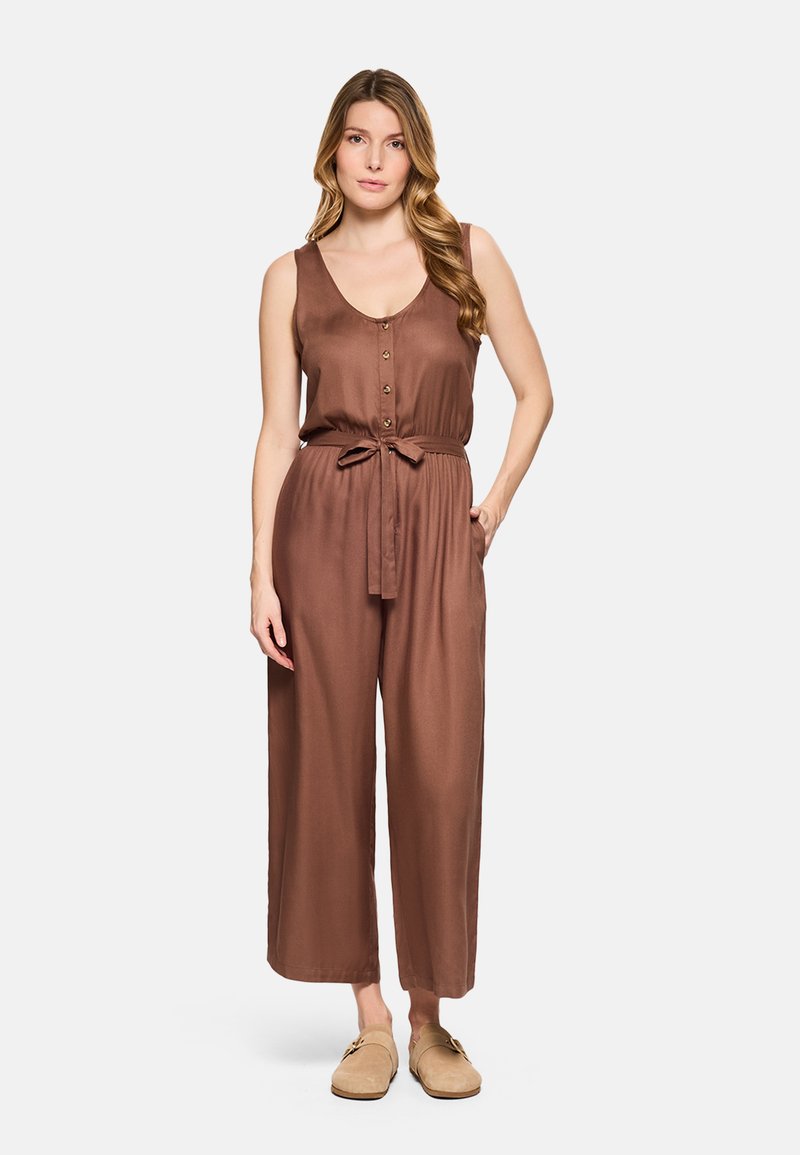 Mujer con cabello largo y ondulado, vestida con un mono marrón sin mangas con botones y cinturón, de pie con una mano en el bolsillo y zapatos beige sin cordones.