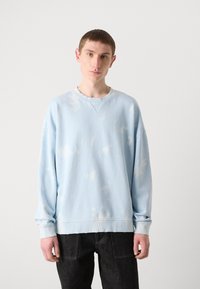 Sudadera azul claro con patrones de tie-dye blancos, hombros caídos, puños y cuello de canalé, combinada con jeans oscuros.