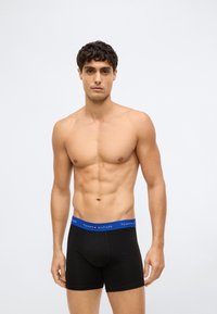 Homem sem camisa com cabelo curto e caracolado, vestindo boxers pretos com uma faixa azul da Tommy Hilfiger, de pé contra um fundo branco simples.