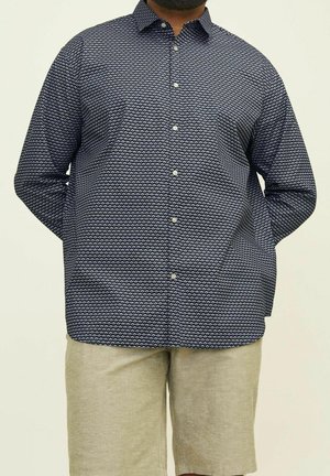 Mężczyzna ubrany w granatową, wzorzystą koszulę na guziki z długim rękawem oraz beżowe szorty, stojący z rękami za plecami.