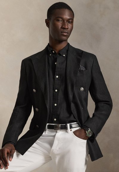 Άνδρας με μαύρο σακάκι διπλής κουμπιάς, μαύρο πουκάμισο με κουμπιά, λευκό παντελόνι, μαύρη ζώνη και ρολόι χειρός, ποζάρει με το ένα χέρι στην τσέπη.