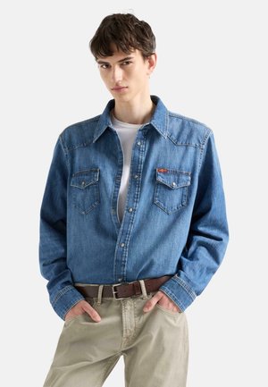 Giovane uomo che indossa una camicia di jeans blu con bottoni sopra una maglietta bianca, pantaloni beige e cintura marrone, in piedi con le mani in tasca.