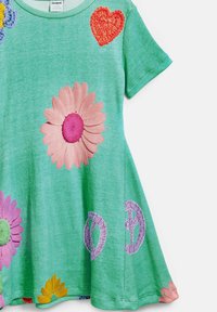 Grön, ribbad klänning med korta ärmar. Har stora broderade blommor och ett hjärta, med fredssymboler i mjuka lila och gula accenter.