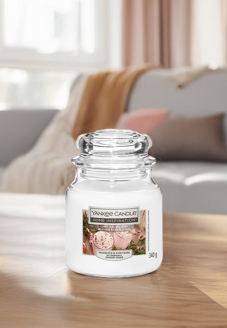 Yankee Candle Home Inspiration Kerzen in der Duftrichtung Zuckerstangen-Milchshake in klarem Glasbehälter auf einem Holztisch, mit verschwommenem Sofa und Kissen im Hintergrund.