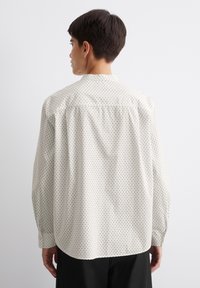 Chemise blanche à pois noirs, à manches longues, avec un col boutonné et une coupe décontractée. Tissu lisse avec un léger éclat, détail de pince au dos.
