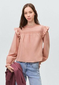 Blusa rosa con trama, maniche a sbuffo, collo rotondo e dettagli arricciati. Abbinata a jeans in denim blu chiaro e tiene una felpa bordeaux.