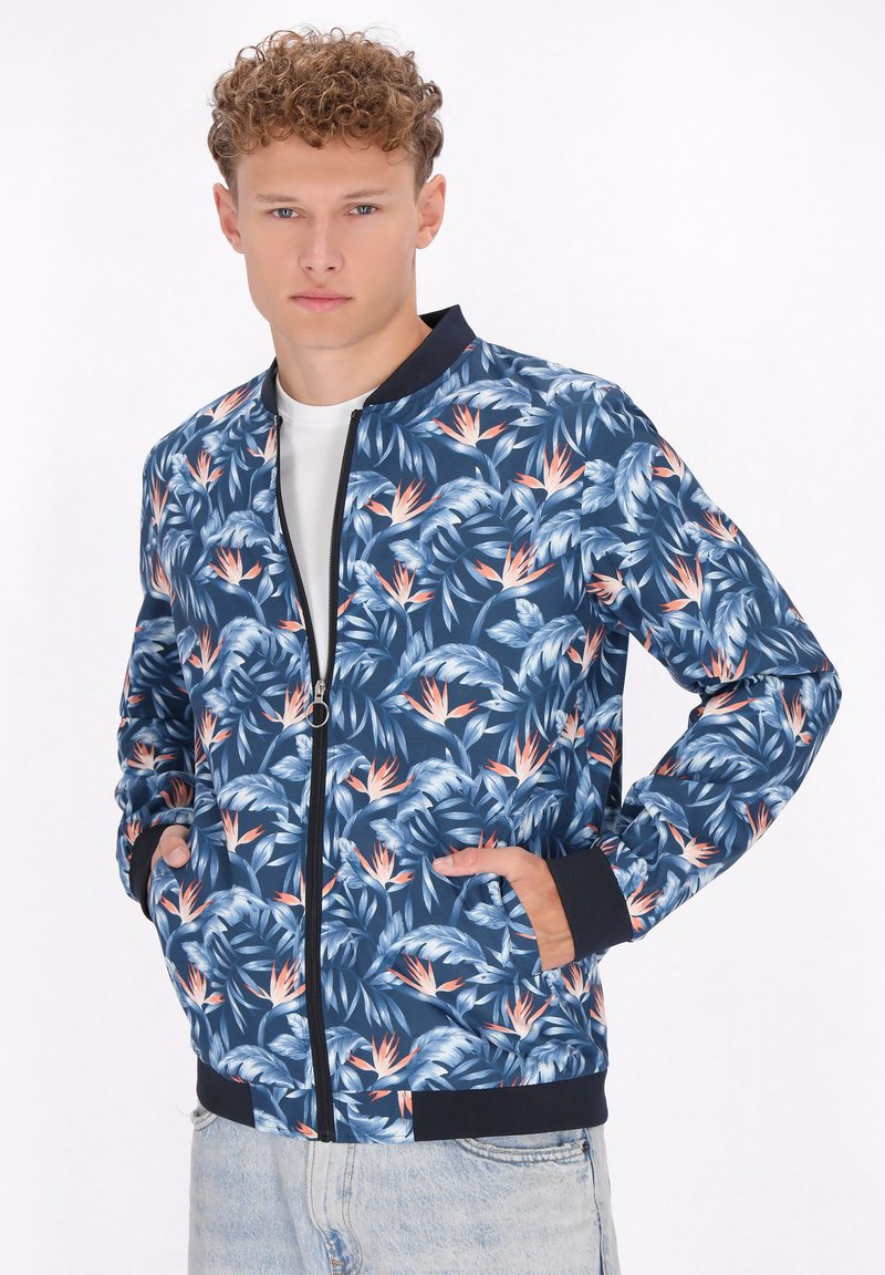 Giacca bomber floreale in marine con dettagli in azzurro chiaro e pesca. Presenta una chiusura con zip, polsini a costine e due tasche laterali. Tessuto leggero.